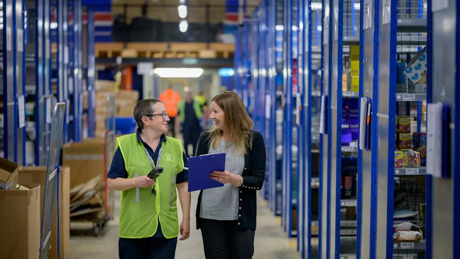 Two colleagues walking down a warehouse aisle with shelving, one holding a handheld scanner