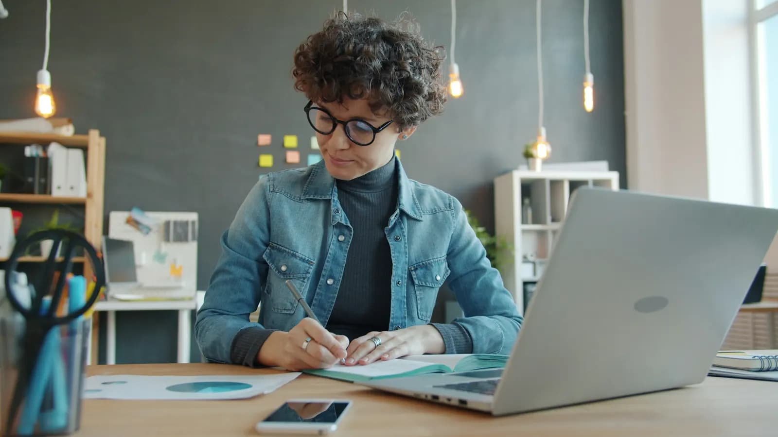 Woman writing in a notebook at a desk beside a laptop